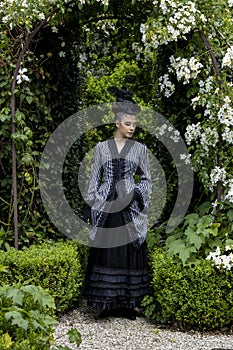 A Victorian woman wearing a striped silk polonaise and a black skirt and standing in a summer garden