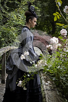 A Victorian woman wearing a striped silk polonaise and a black skirt and standing in a summer garden