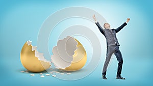 A victorious businessman stands near a large broken golden eggshell on blue background.