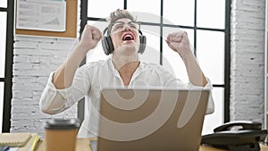 Victorious young caucasian man celebrating winning business success at office, exuding confidence with laptop and headphones