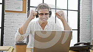 Victorious young caucasian man celebrating winning business success at office, exuding confidence with laptop and headphones