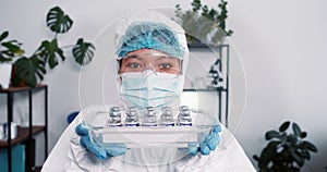 Victory over coronavirus. Cheerful female science lab worker in protection suit shows medical tray with vaccine flasks.