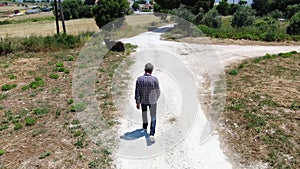Video from above of a man wearing jeans, a plaid shirt and sunglasses man walking along a country road. Dron pursues