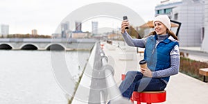 Video call. Adult smiling woman in knit cap sits with coffee on the city embankment and using her mobile, format photo