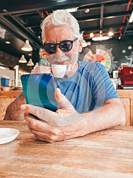 Video call concept. Handsome smiling bearded senior man drinking a coffee cup sitting at cafe table talking on mobile phone using