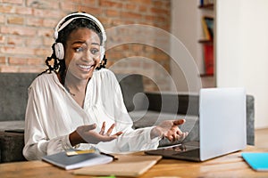 Video call. Emotional black woman in headphones talking and gesturing at laptop webcamera, sitting at home