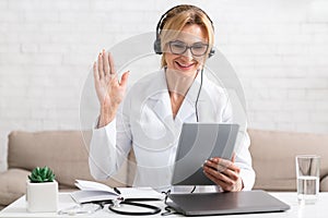 Video call to therapist. Smiling woman doctor in headphones greets the patient and holds at tablet