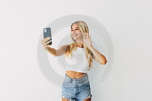 Video communication via smartphone. A young woman is using phone for video call, online meeting, standing against white background