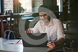 Video conferention. Shows greetings gesture. Adult man sits in cafe at daytime and using the laptop for the remote work