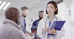 Video of diverse male patient and female doctor with clipboard talking in busy hospital corridor