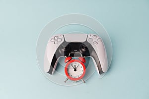 Video game console gamepad on blue background with red alarm clock. Tim to play. Gamer problem in dependance of gaming.