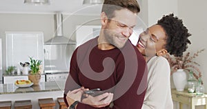 Video of happy diverse couple hugging and using smartphone in kitchen