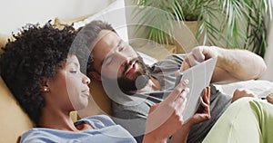 Video of happy diverse couple relaxing at home, lying on bed using tablet