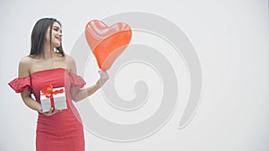 Video of joyful woman holding a gift box and heart balloon, smiling over a white background.