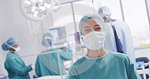 Video portrait of biracial female surgeon in mask smiling in operating theatre, copy space