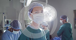 Video portrait of caucasian female surgeon in face mask smiling in operating theatre, copy space