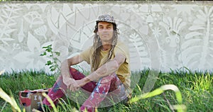 Video portrait of smiling caucasian man with dreadlocks sitting on grass by mural on wall