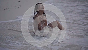 Video of smiling girl sitting on the beach and playing with wave splash in slow motion.