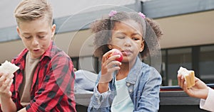 Video of three diverse schoolchildren eating packed lunches, talking in schoolyard