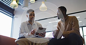 Video of two diverse businessmen talking and using tablet in lounge area of busy office