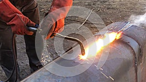 Video view of the welder through the fire. A welder in protective clothing in the open air cuts metal pipes with