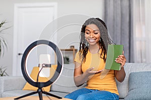 Videoblog and technology concept. Positive black female blogger recording video of book review at home