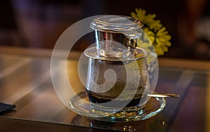 Vietnamese coffee pot sits on a wooden table, with bright yellow flowers in the background