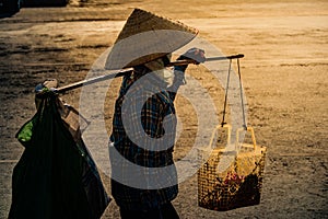 Vietnamese with conical hat carries a yoke on her shoulder along the street.