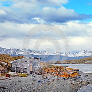 View of an abandoned house and mountains from an island covered by brown and orange vegetation on the Beagle Channel