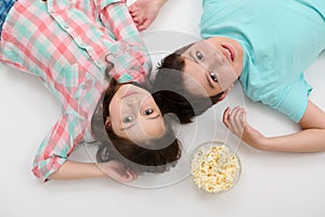 View from above of adorable kids in casual clothes lying on white background with a bowl of popcorn, looking at camera