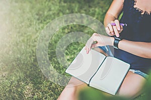 View from above. Close-up of notepad lying on girl`s lap.Girl sitting in garden on lawn under tree uses smartwatch