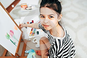 View from above of a creative little girl kid smiling, looking at the camera during painting on paper on an easel at home. A happy