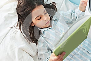View from above of a cute little girl lying in the bed and reading the fairy tale book. Kid in pajamas relaxing and reading