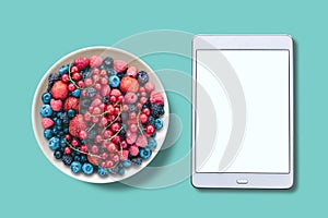View from above. Different berries in a plate. Near the tablet with an assorted white screen.