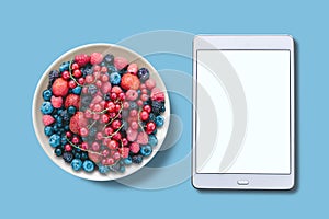 View from above. Different berries in a plate. Near the tablet with an assorted white screen.