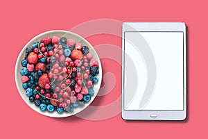 View from above. Different berries in a plate. Near the tablet with an assorted white screen.