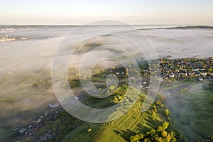 View from above of foggy green hills, village house roofs in valley among green trees under bright blue sky. Spring misty