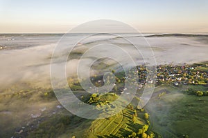 View from above of foggy green hills, village house roofs in valley among green trees under bright blue sky. Spring misty