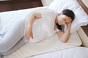 View from above of a multi ethnic beautiful adult pregnant woman holds hand on belly, sleeping in a light bedchamber