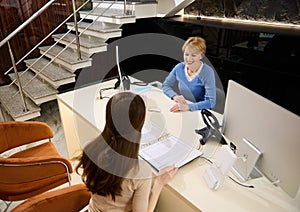 View from above to pleasant mature European woman retail manager, consultant sitting at reception desk in modern office building