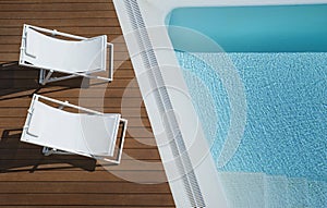 View from above of two white canvas chaise lounge chairs positioned on a brown outdoors wooden deck by a turquoise infinity pool