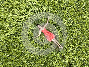 View from above of young attractive slim woman in red dress laying with closed eyes in green wheat field