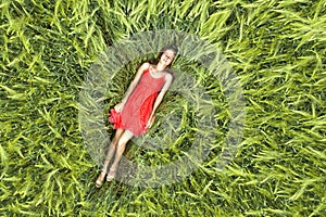 View from above of young attractive slim woman in red dress laying with closed eyes in green wheat field