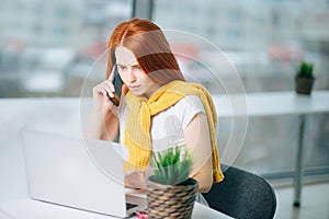 Business woman sitting in office at desk, looking laptop and uses smartphone