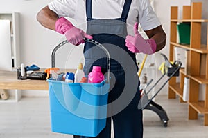 View of African american cleaner showing thumb up and holding bucket of detergents in office