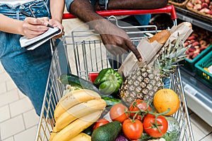 View of african american man pointing with finger at shopping cart with groceries near woman with notebook