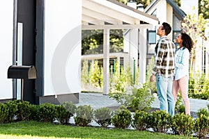 View of african american man and woman looking at house while standing near lawn