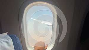 View of the airplane window with a hand on it and the wing of the aircraft and the sky