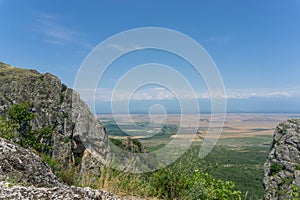 View of the Alazani Valley from a high mountain. Rocks and trees around. Agrarian fields are visible. Clear blue sky and clouds