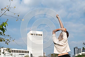 View from the back. Asian woman wearing white shirt is in a cooldown posture after exercising in the park. The background is part